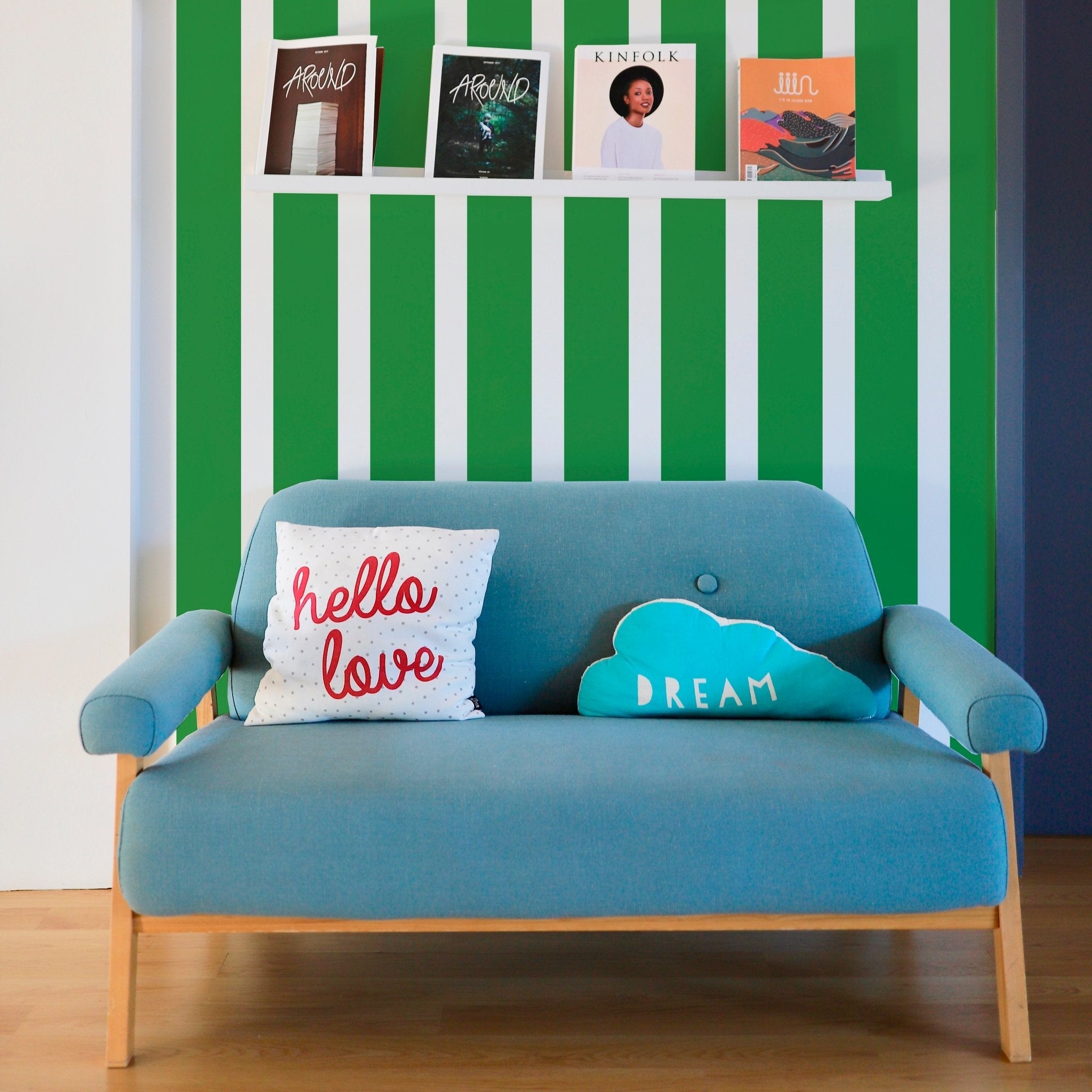 Traditional wallpaper in British green stripe shown behind a colorful blue love seat and book shelving
