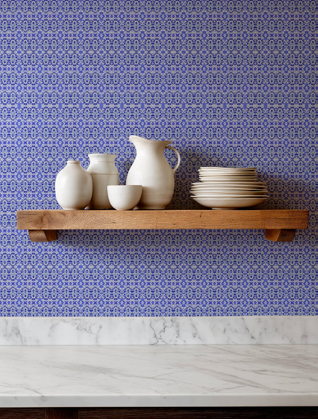 A wooden floating shelf mounted on a blue and white patterned wallpaper wall, holding a collection of matte white ceramic vases, a pitcher, and stacked plates, with a white marble countertop below.